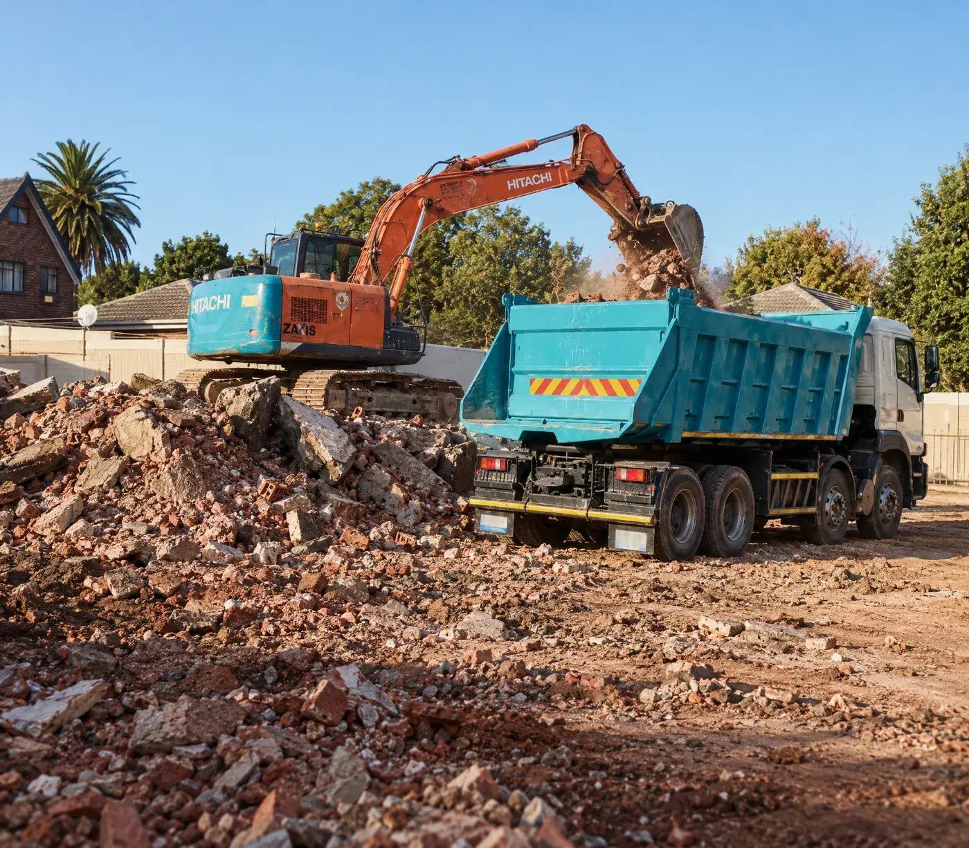 Hitachi excavator tipping demolition rubble into a teal tipper truck on a cleared suburban site in Cape Town