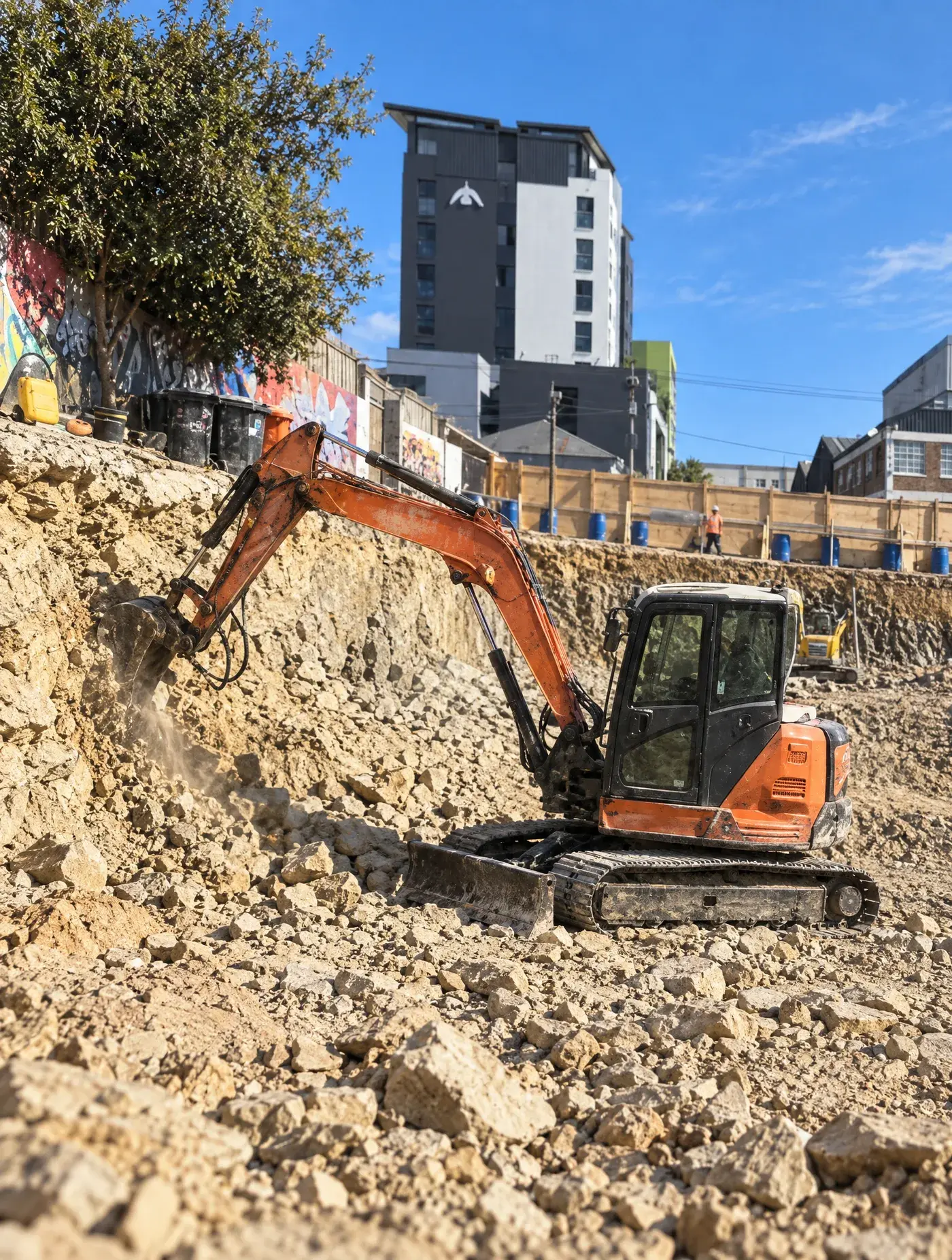 Orange mini excavator digging out a rock basement excavation between city buildings