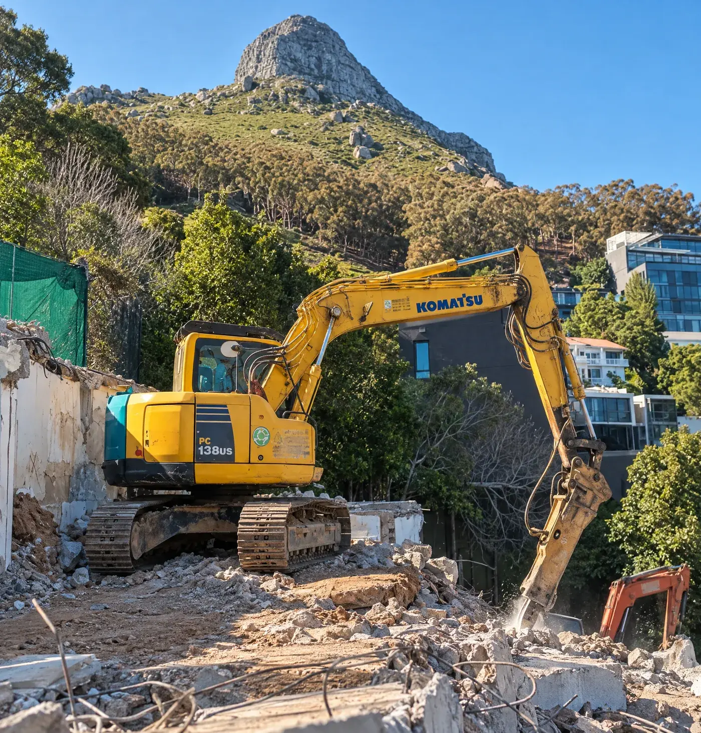 Rubble Cycle Komatsu PC138US excavator with a hydraulic breaker breaking concrete on a demolition site below Lion's Head