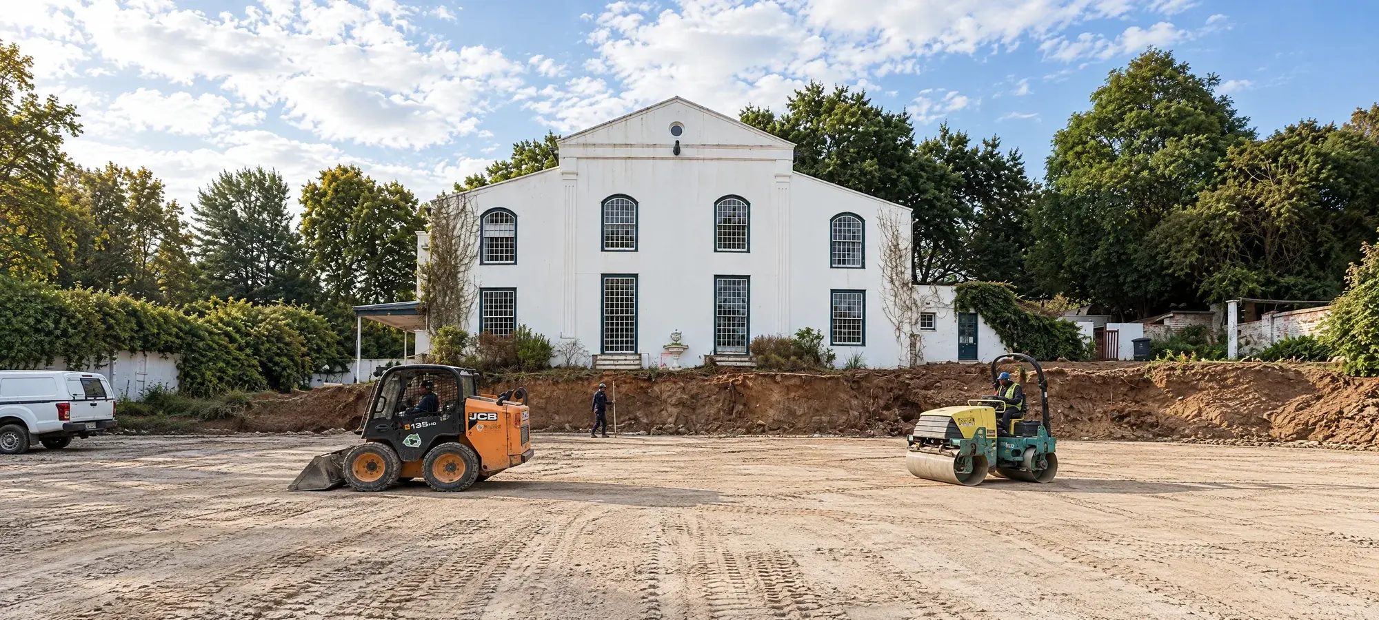 Skid steer loader and roller levelling a cleared site in front of a white heritage building