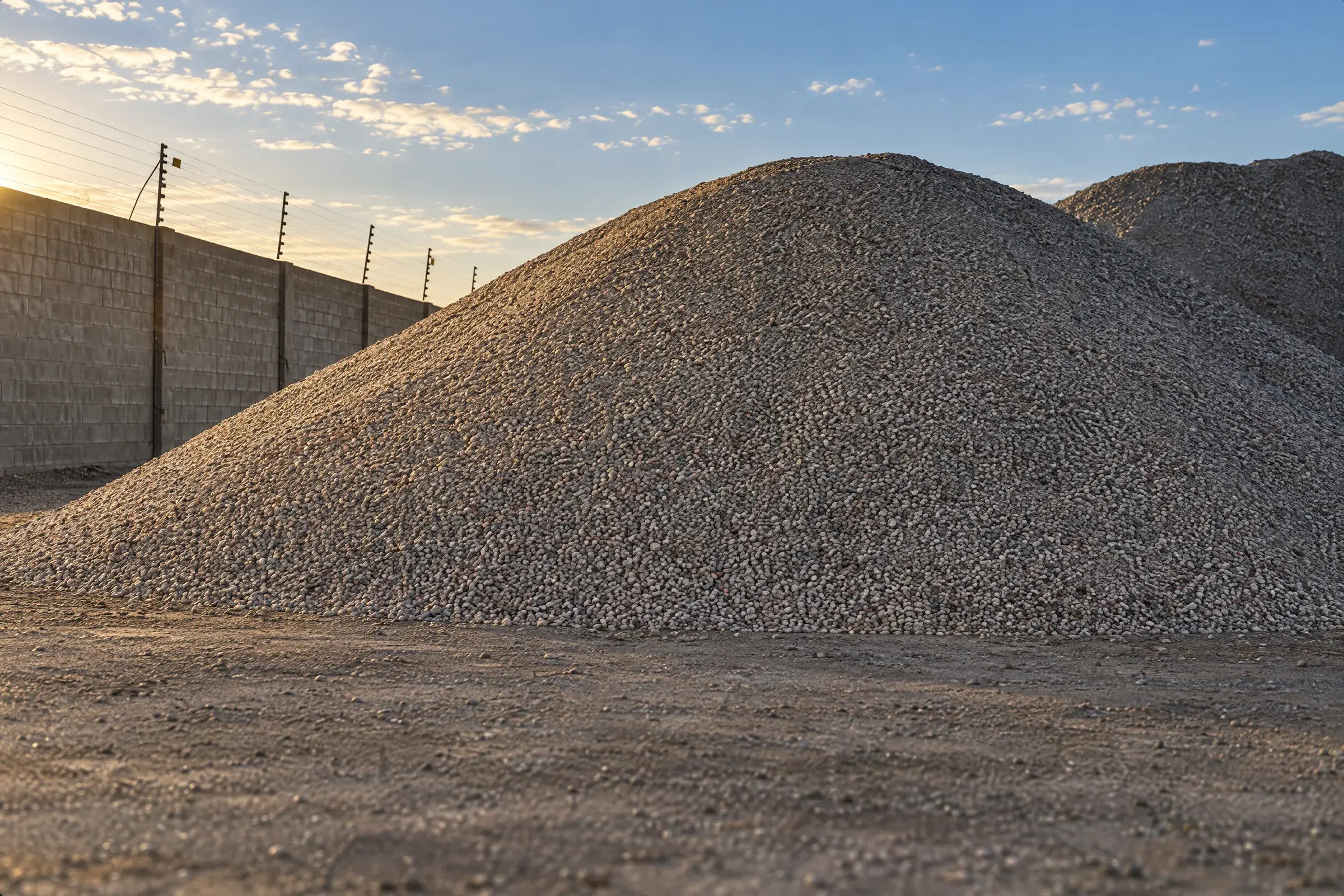 Conical stockpile of fine crushed recycled aggregate against a block wall with an electric fence at sunset
